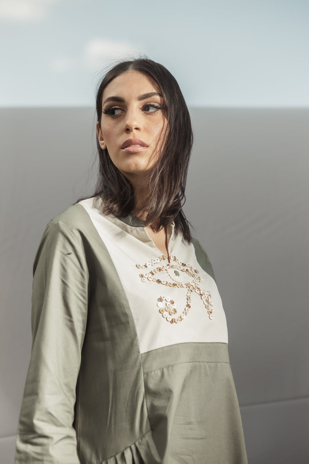 A long sleeve poplin cotton kaftan in khaki and beige consisting of a miniature V-neckline and an ankle length relaxed fit detail. Embellished by hand embroidered design of the Eye of Horus showcasing unique hand embroidery through stones of shades of beige completed by green beads, finished by hand stitched thread. The Eye of Horus is an Ancient Egyptian symbol of protection, royal power and good health.